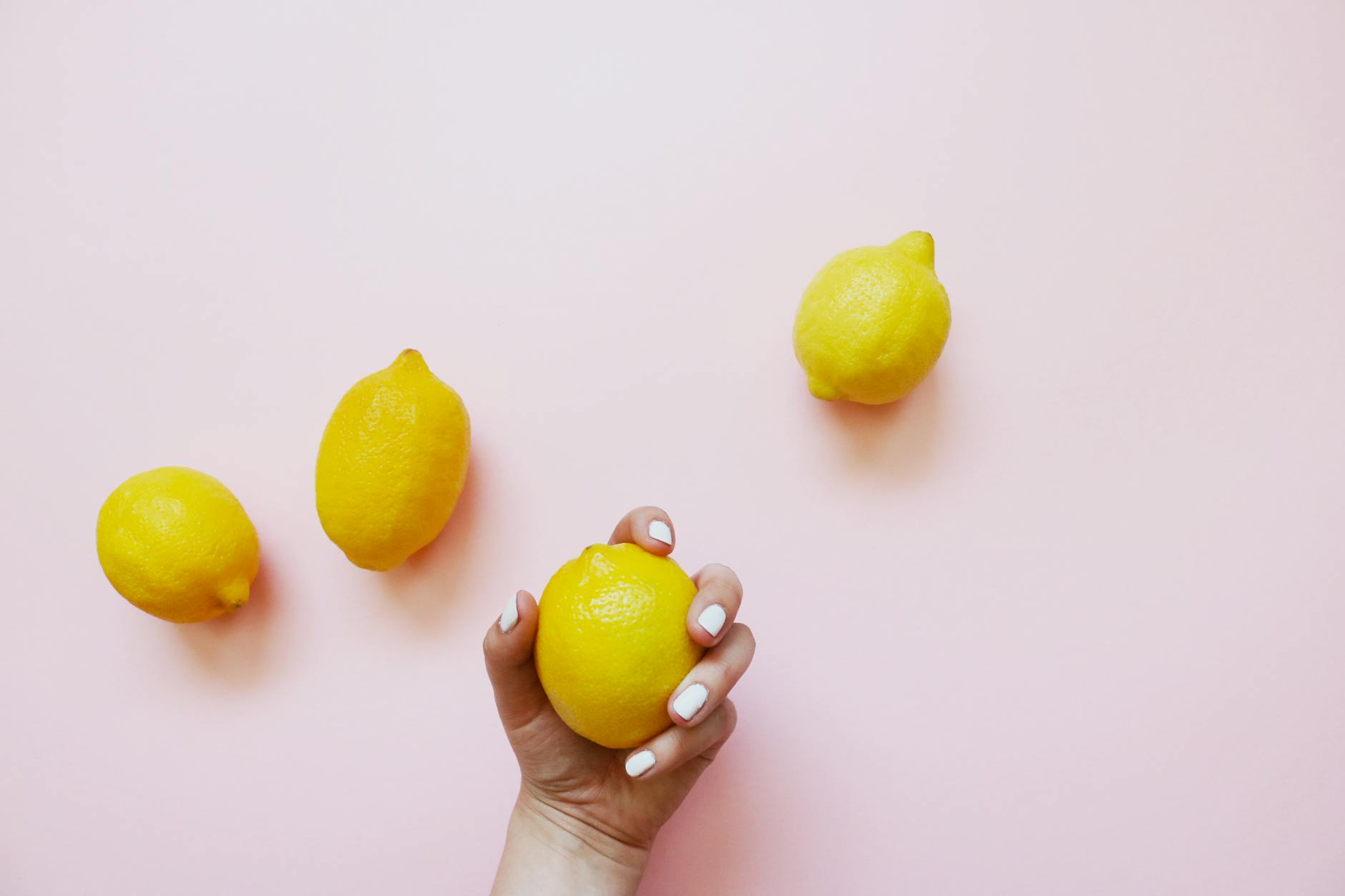 A hand with white nails holding a lemon on a soft pink background, surrounded by three additional lemons