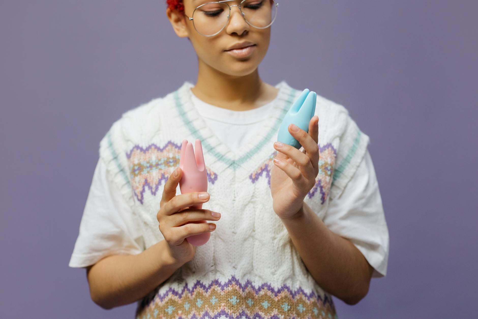 Woman holding blue and pink silicone vibrators while wearing eyeglasses, exploring personal pleasure.